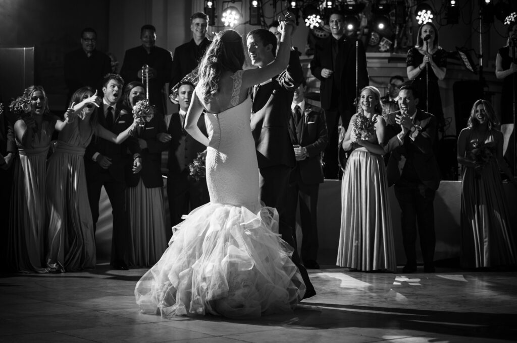 Bride and groom sharing their first dance at a Please Touch Museum wedding reception, surrounded by applauding guests and a live band in the background, in black and white.