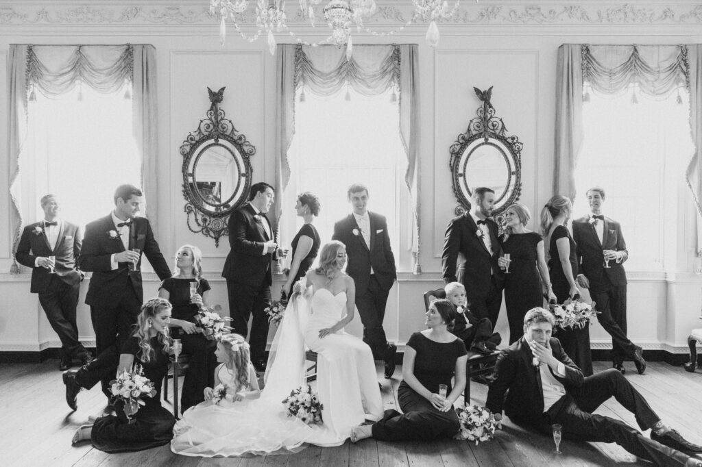 Black and white photo of a Powell House Philadelphia wedding: bride seated, groom standing behind her, surrounded by bridesmaids and groomsmen in an elegant room with mirrors.