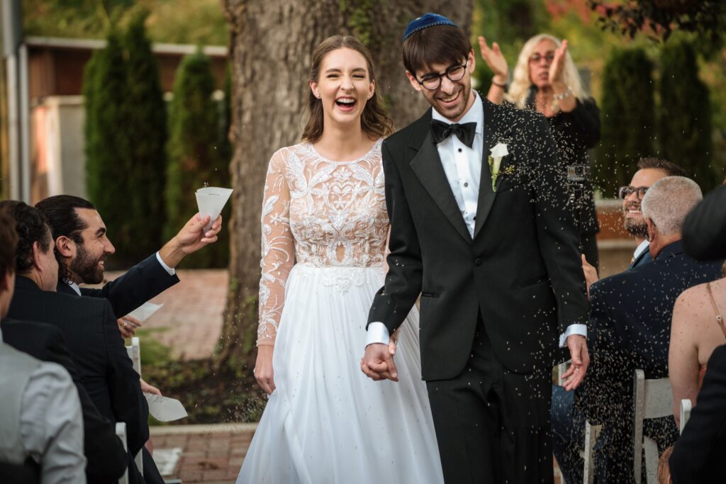 A joyful bride and groom holding hands, laughing while walking down the aisle as guests throw rice at them during a Hotel du Village wedding ceremony.