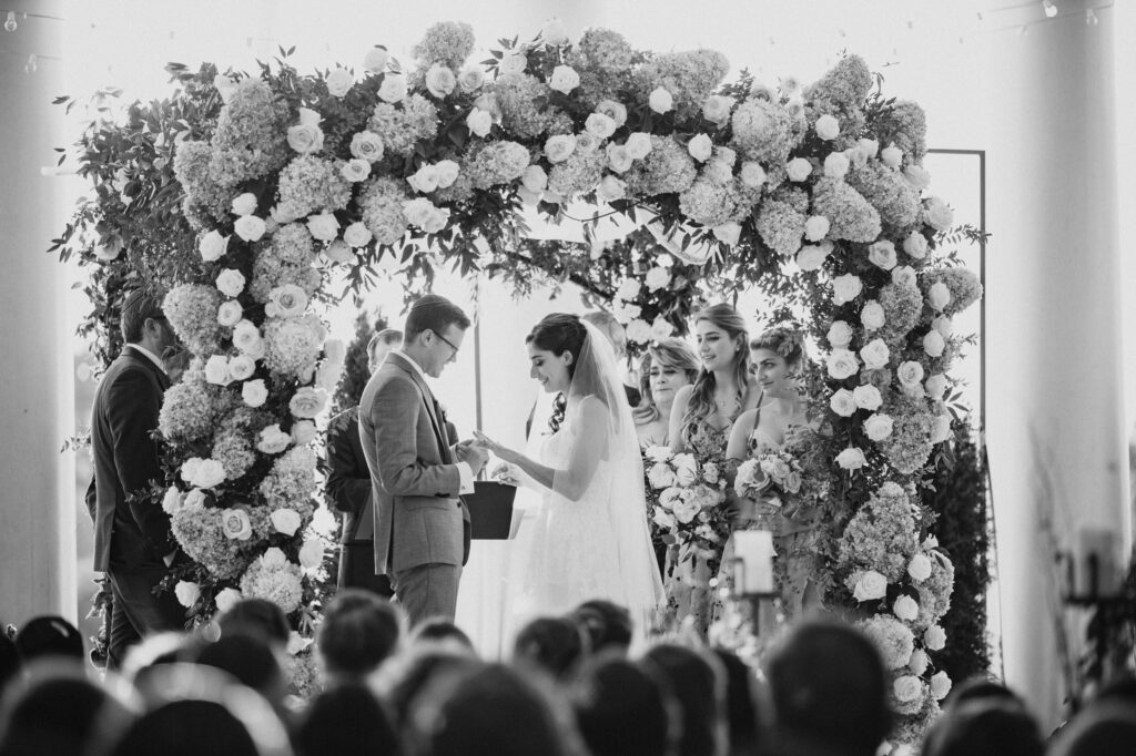 A bride and groom exchange vows under a floral arch at the Water Works in Philadelphia, surrounded by guests and bridesmaids during an outdoor wedding ceremony.