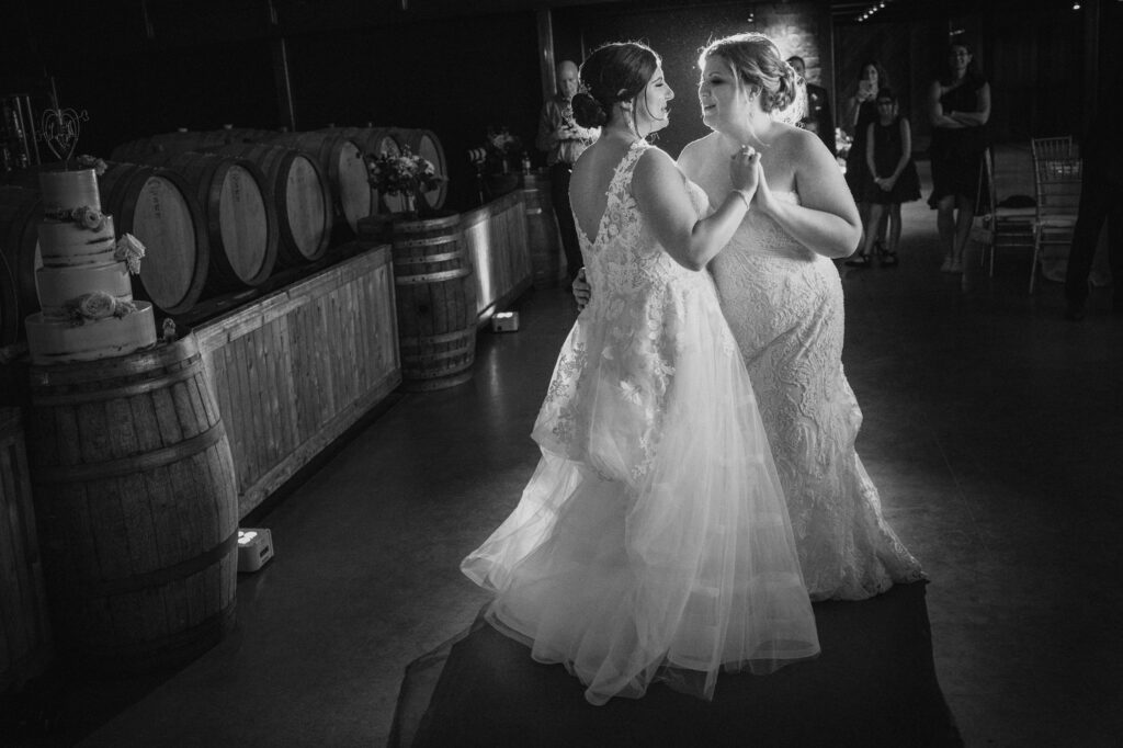 Two brides in elegant white gowns share a tender moment at Saltwater Farms Vineyard, standing close together in a dimly lit room with wooden barrels and a wedding cake in the background.