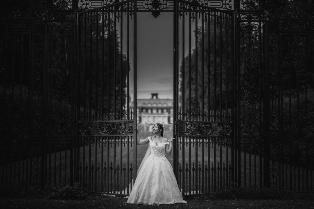 Bride standing at an open gate leading to Old Westbury Gardens, a grand building, at night.