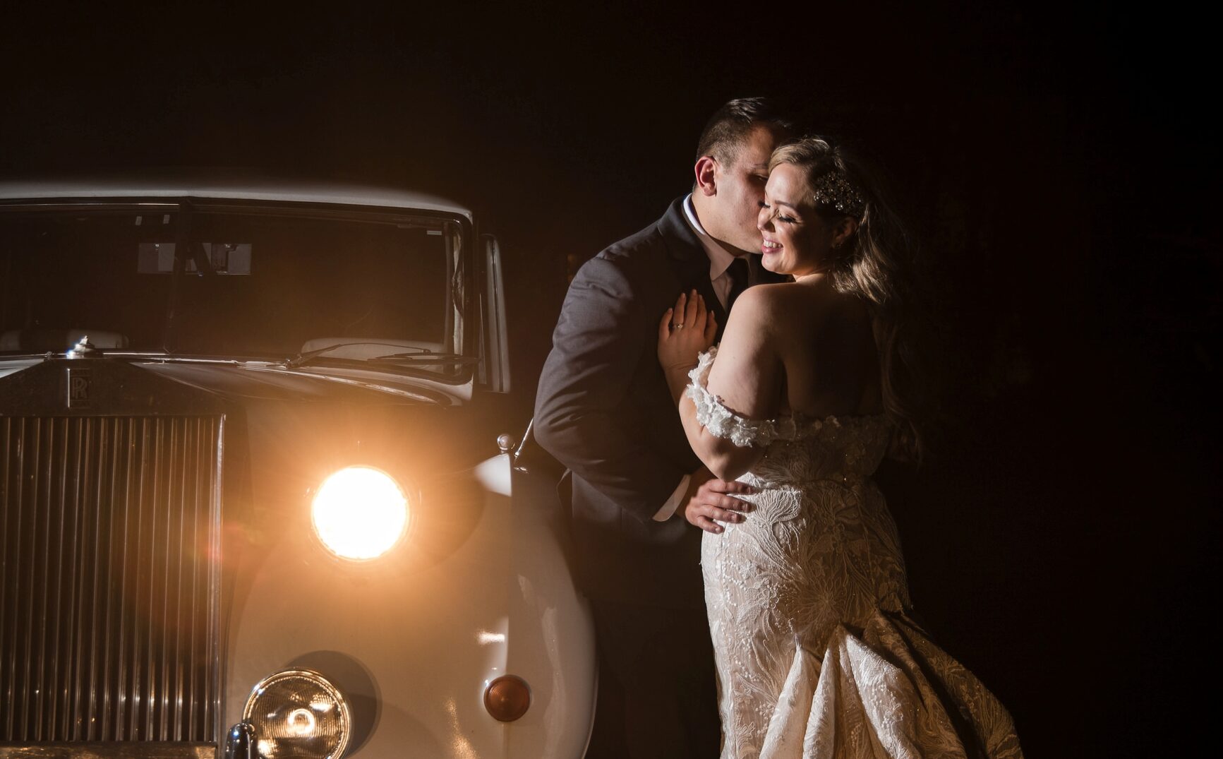 A couple embraces and kisses beside a vintage car with headlights on in a dimly lit setting at their Rock Island Lake Club wedding.