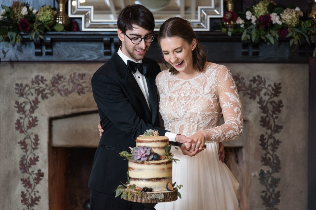 A bride and groom in formal attire cutting a three-tiered wedding cake together, smiling, at a Hotel du Village wedding, with an ornate fireplace and floral decorations in the background.