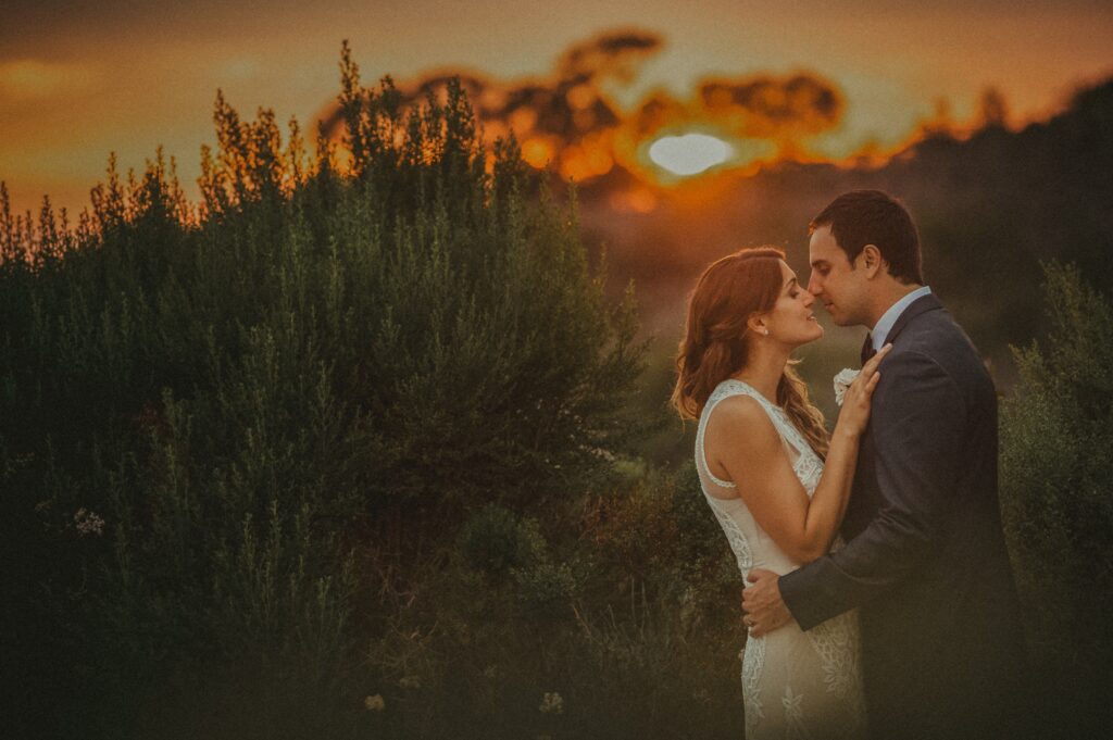 A couple in wedding attire embraces at sunset during their Resort at Pelican Hill wedding, surrounded by lush greenery under a glowing orange sky.