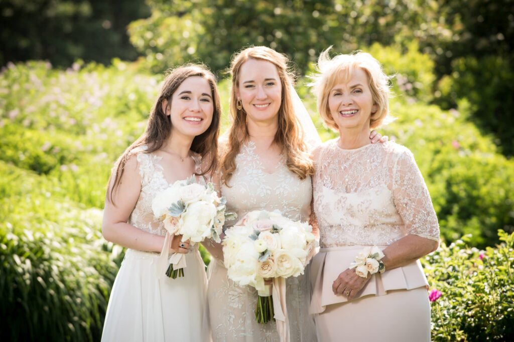 Three smiling women dressed in formal attire with bouquets posing together outdoors at an ocean house Rhode Island wedding.