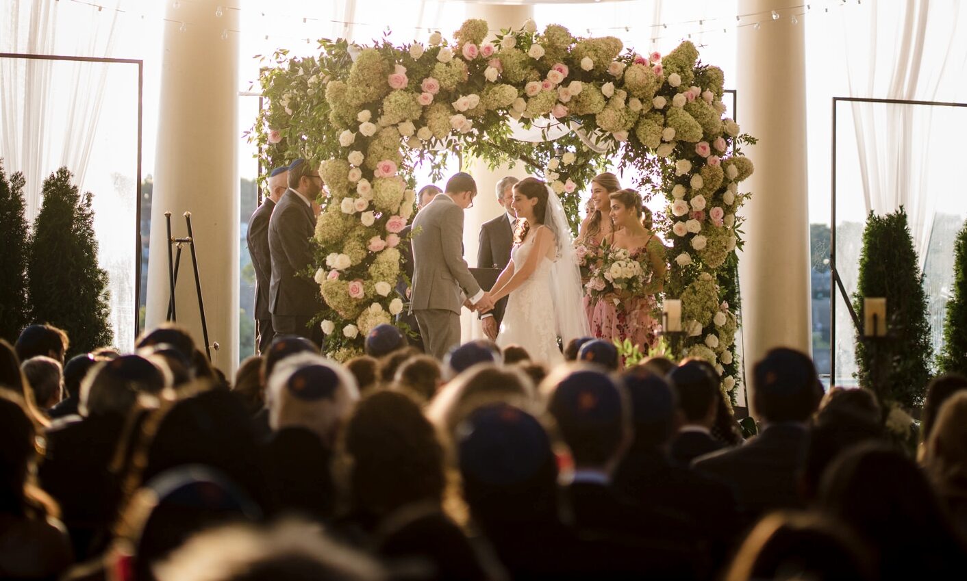 A Water Works Philadelphia wedding ceremony in a large room with a floral arch, bride and groom facing an officiant, and guests seated in the foreground.
