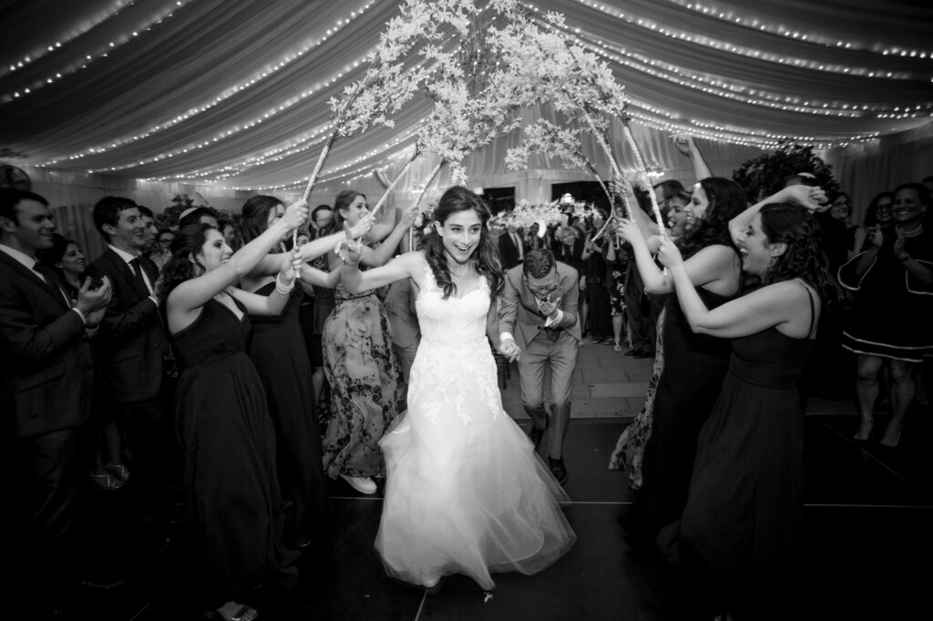 Bride smiling and dancing under a floral chandelier at her Water Works Philadelphia wedding reception, surrounded by cheering guests.