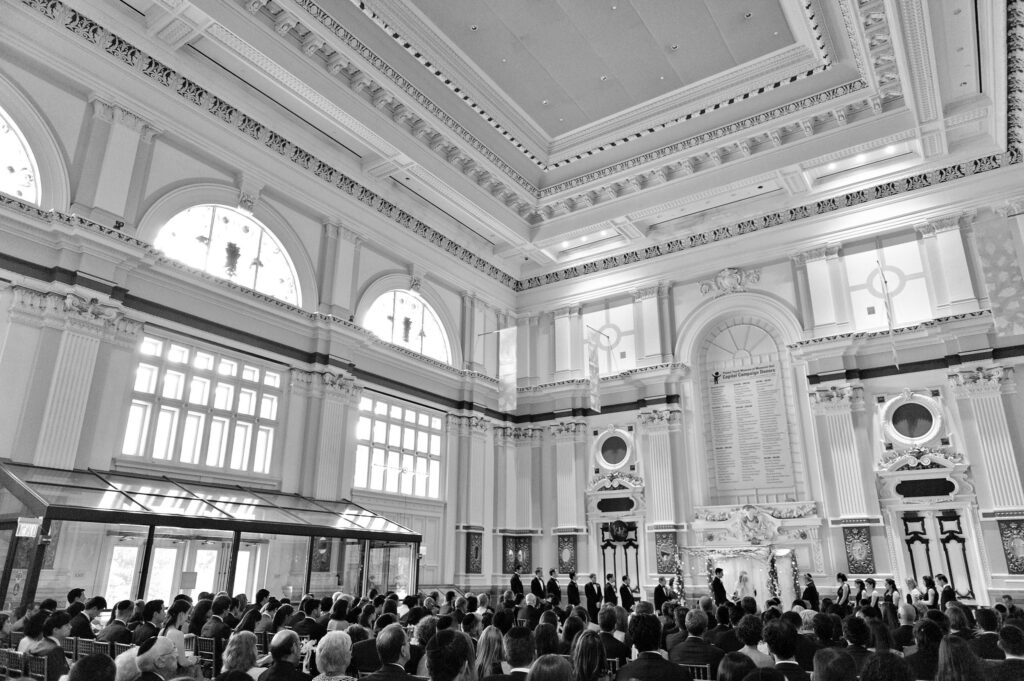 A black and white photo of a Please Touch Museum wedding ceremony, featuring a grand hall with large windows and an audience.