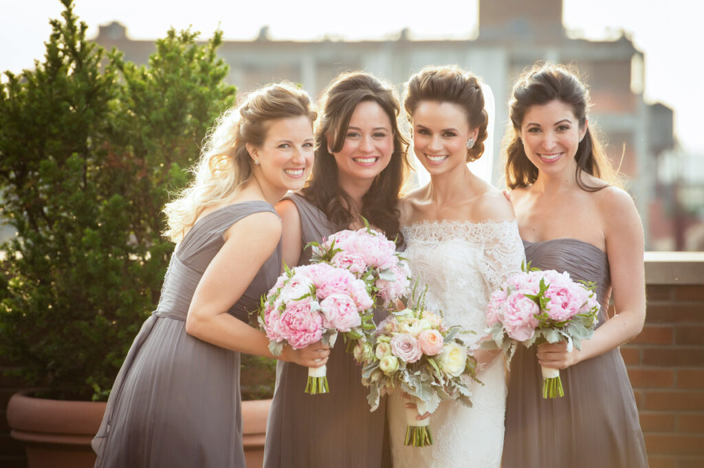 Joyful bridal party posing on a rooftop, with bridesmaids in gray dresses holding bouquets of pink peonies, and the bride in a lace gown, smiling widely