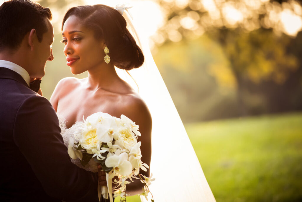 Elegant bride in a strapless gown with a white floral bouquet, gazing lovingly at her groom in a twilight park setting, showcasing a serene atmosphere