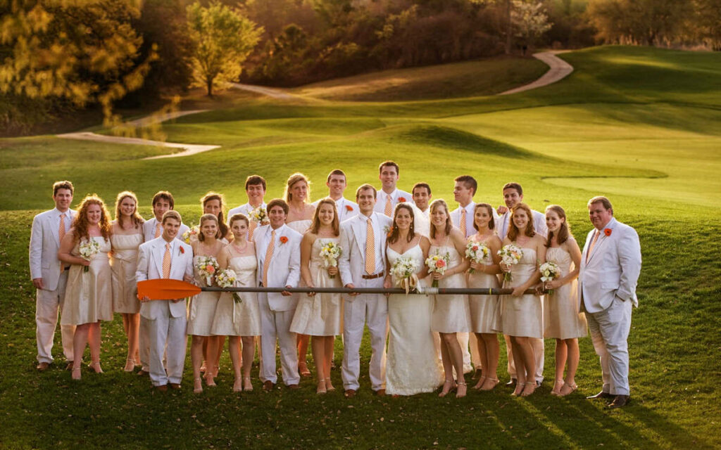 Large wedding party standing on a lush golf course, with bridesmaids in cream dresses and groomsmen in white suits with orange accents, embodying a chic, coordinated look