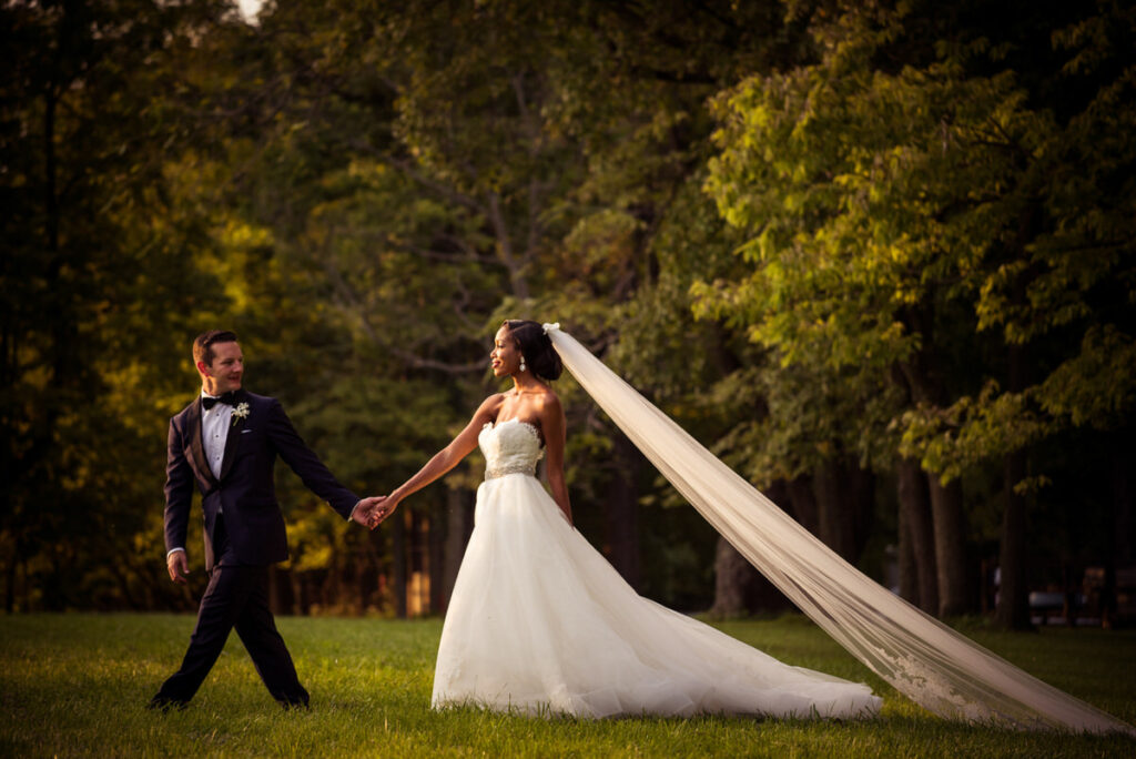 Bride in a stunning white gown with a long flowing veil and groom in a classic tuxedo, walking hand in hand in a peaceful wooded area, exuding happiness