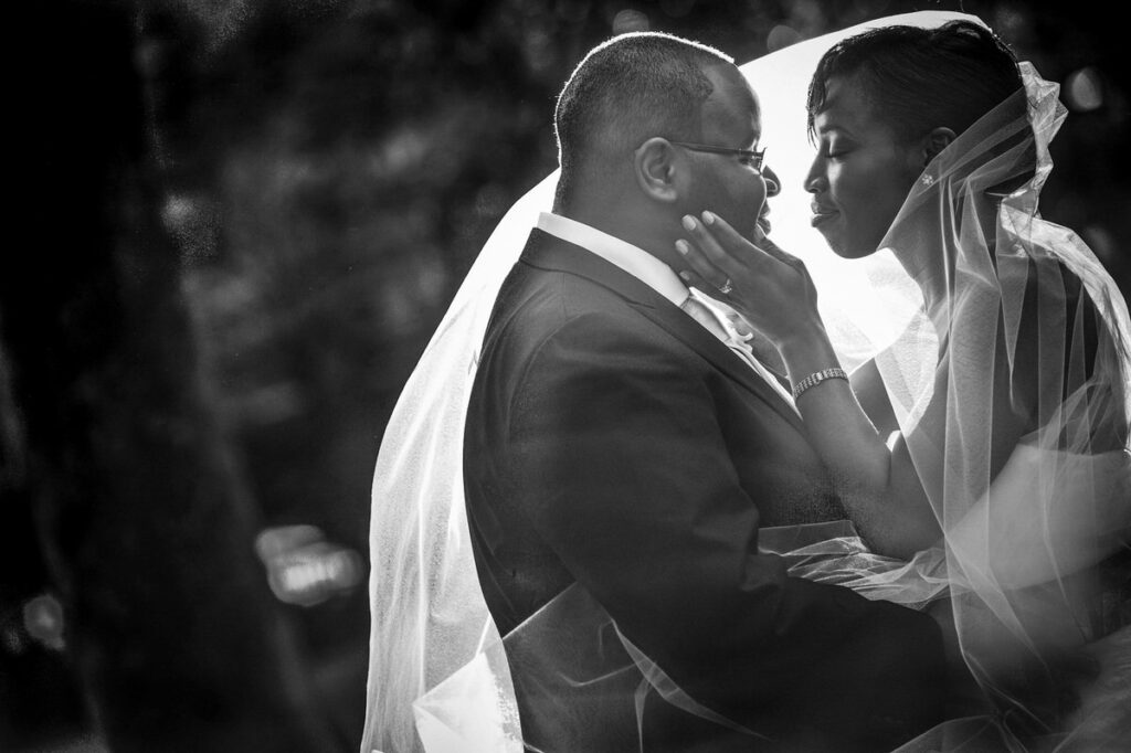 Black and white photo of a tender moment where a groom in a tuxedo gently holds his bride's face under her sheer veil, both sharing a serene expression
