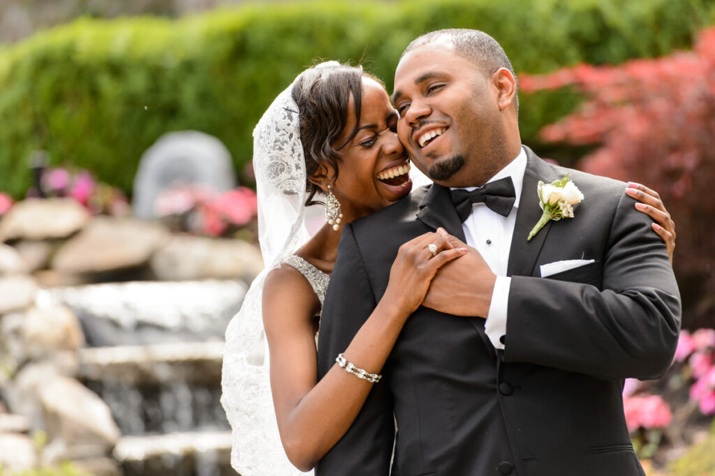 Radiant couple laughing together, the bride in a lace-detailed gown and the groom in a sleek black tuxedo with a boutonniere, against a vibrant floral backdrop