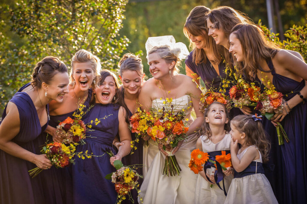 Bridal party captured in a candid moment, with bridesmaids in deep blue dresses and the bride in a strapless gown, all laughing and surrounded by autumnal flowers