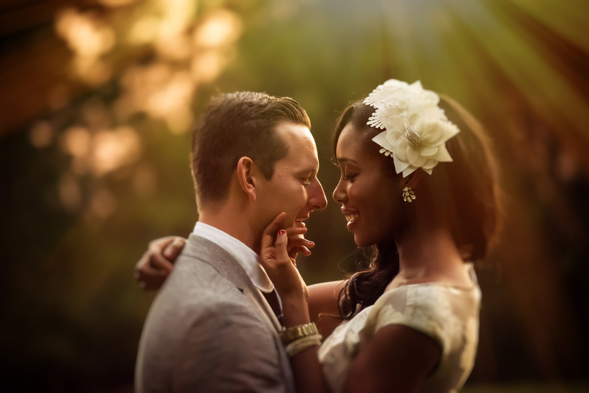 Intimate moment of a diverse couple with the groom in a light suit and the bride wearing a white dress with a floral hairpiece, basking in the golden sunlight