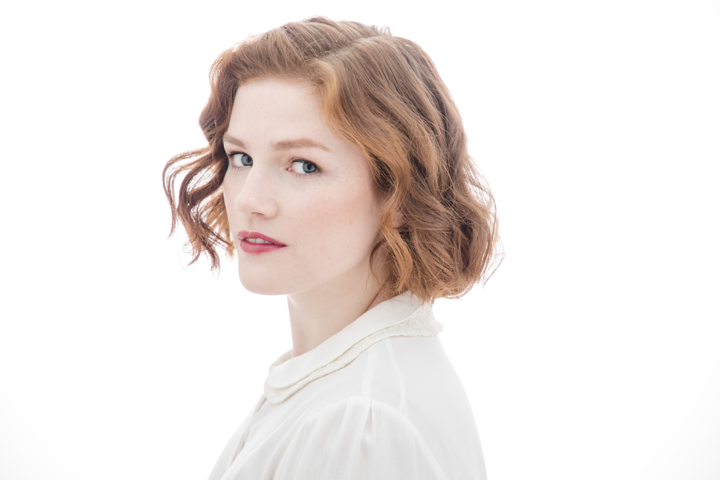 A person with short, wavy red hair and light skin looks over their shoulder, wearing a white shirt against a plain white background, captured perfectly by a renowned New York headshot photographer.