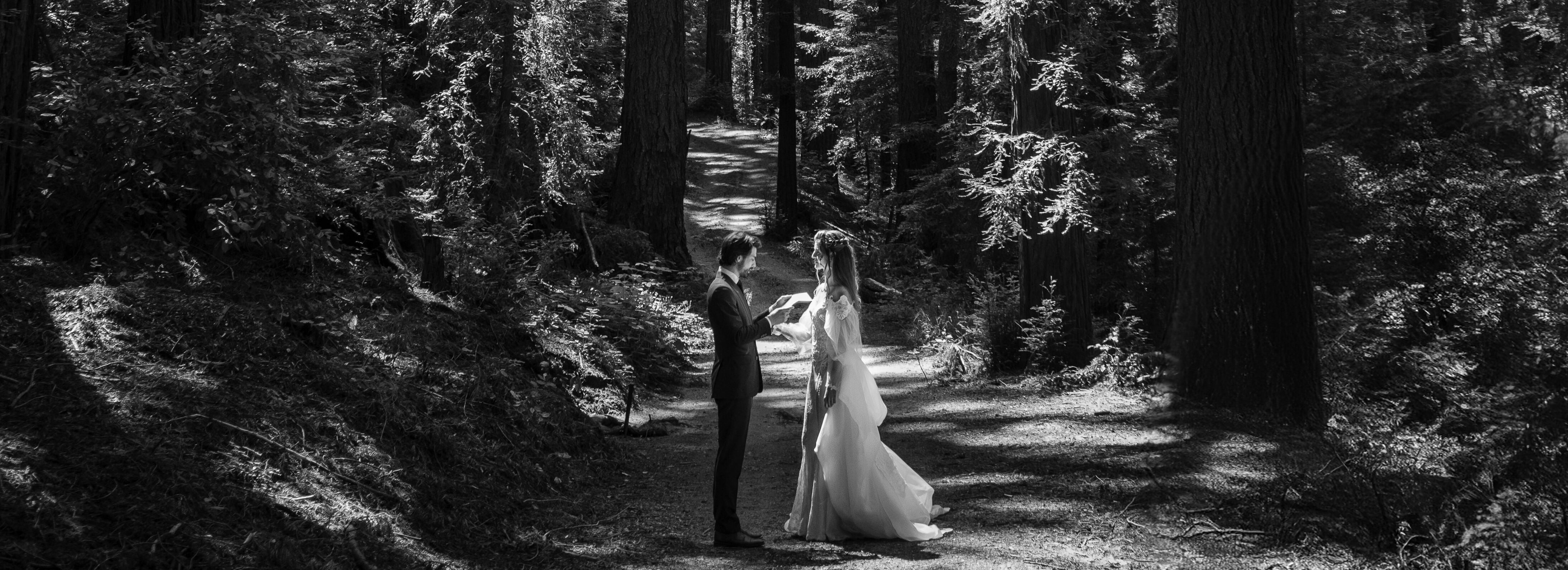 A bride and groom stand facing each other in a sun-dappled forest path at their Nestldown Wedding, holding a piece of paper.