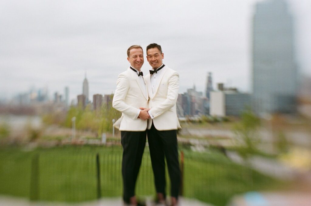 A film wedding photo of two grooms holding hands with a city skyline in the distance