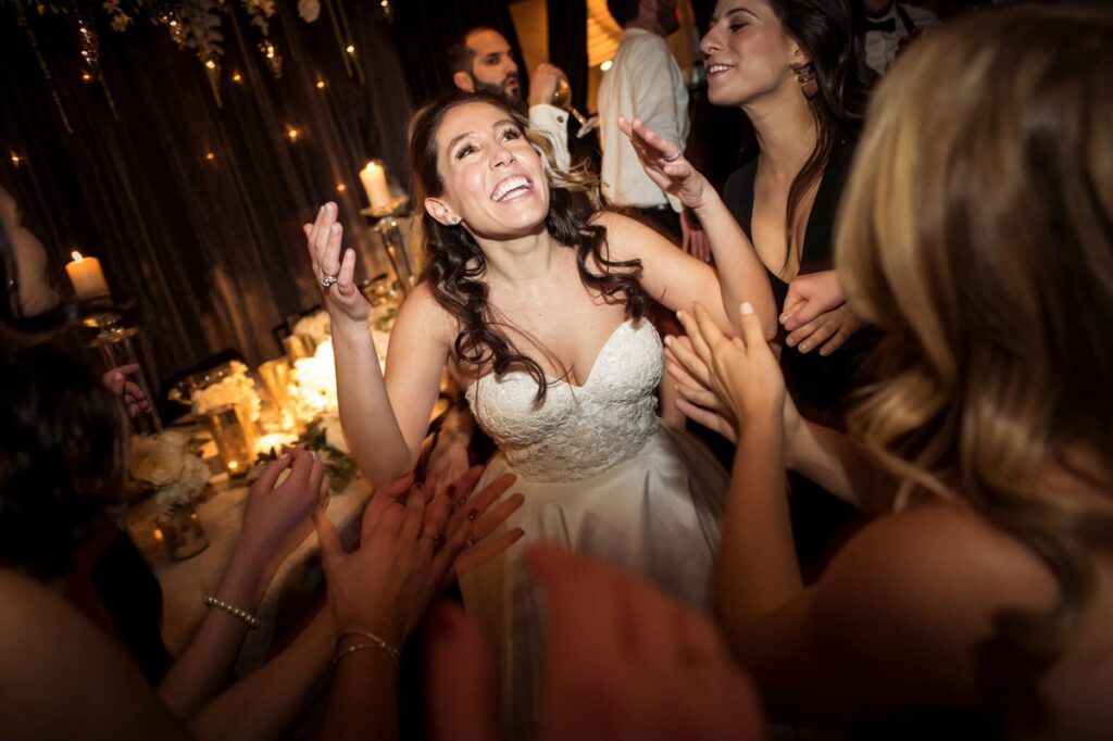 A joyful bride surrounded by her friends claps and smiles broadly in a candlelit reception area, capturing a moment of excitement and celebration at her wedding.
