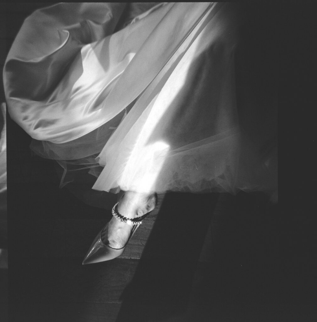 A detailed black-and-white photo focuses on a bride’s foot peeking out from her flowing dress, adorned with a decorative anklet, capturing the elegance and anticipation of the wedding day.