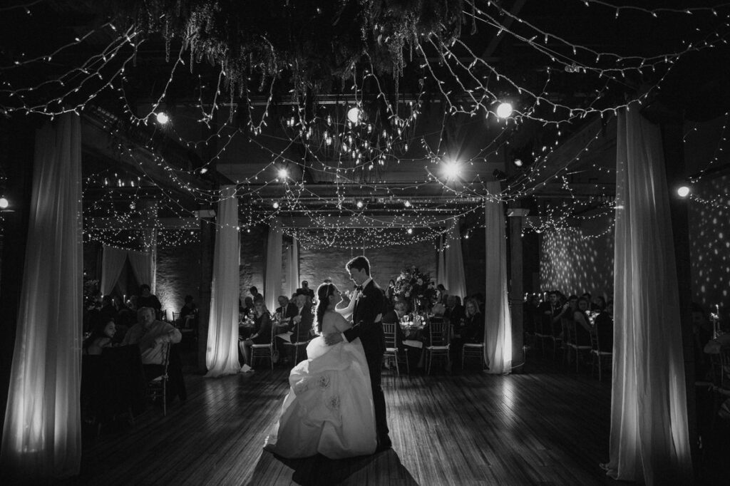 A bride and groom dance under twinkling lights and draped foliage in a magical, dimly lit reception hall, surrounded by guests and exuding a romantic, enchanting atmosphere.