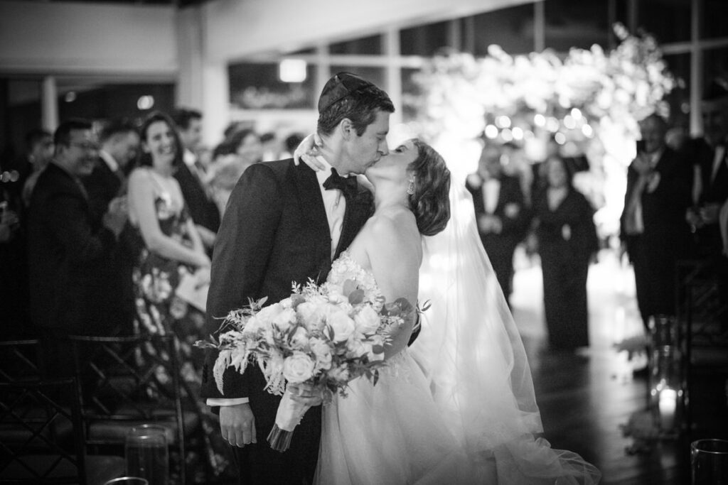 A romantic black-and-white shot of a bride and groom kissing at their wedding reception, surrounded by guests and beneath elegant floral arrangements, conveying a timeless wedding moment.