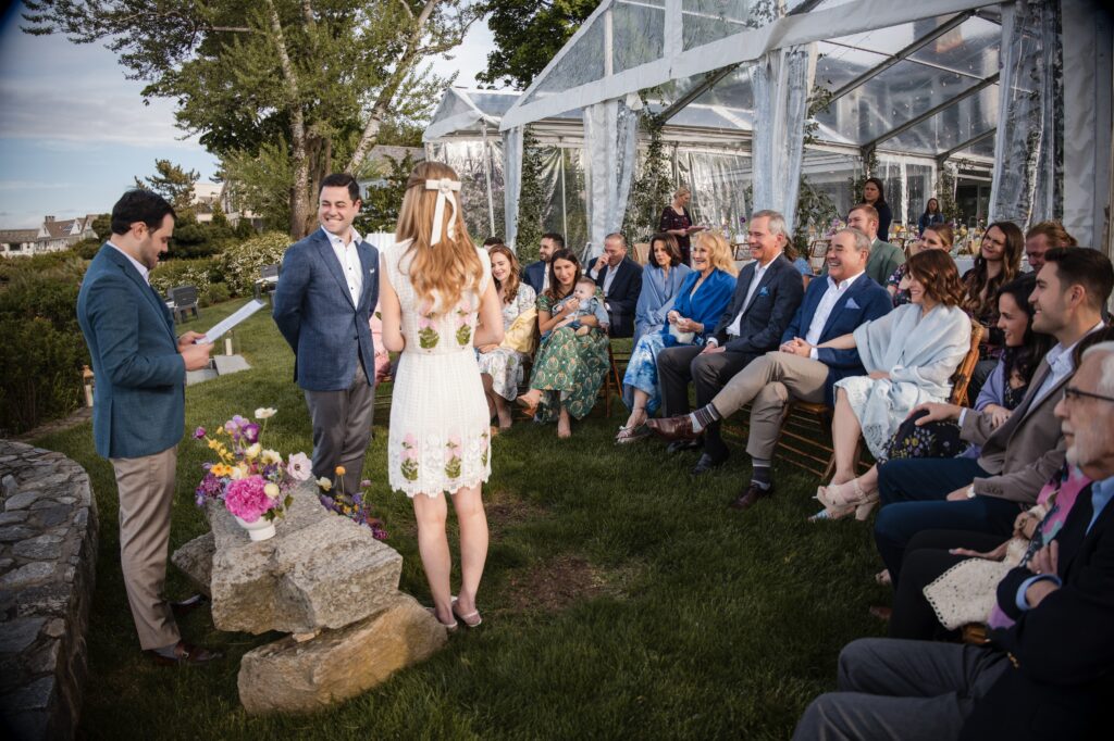 A couple stands at an outdoor wedding ceremony in Westport, Connecticut, exchanging vows under a transparent tent with guests seated beside them.