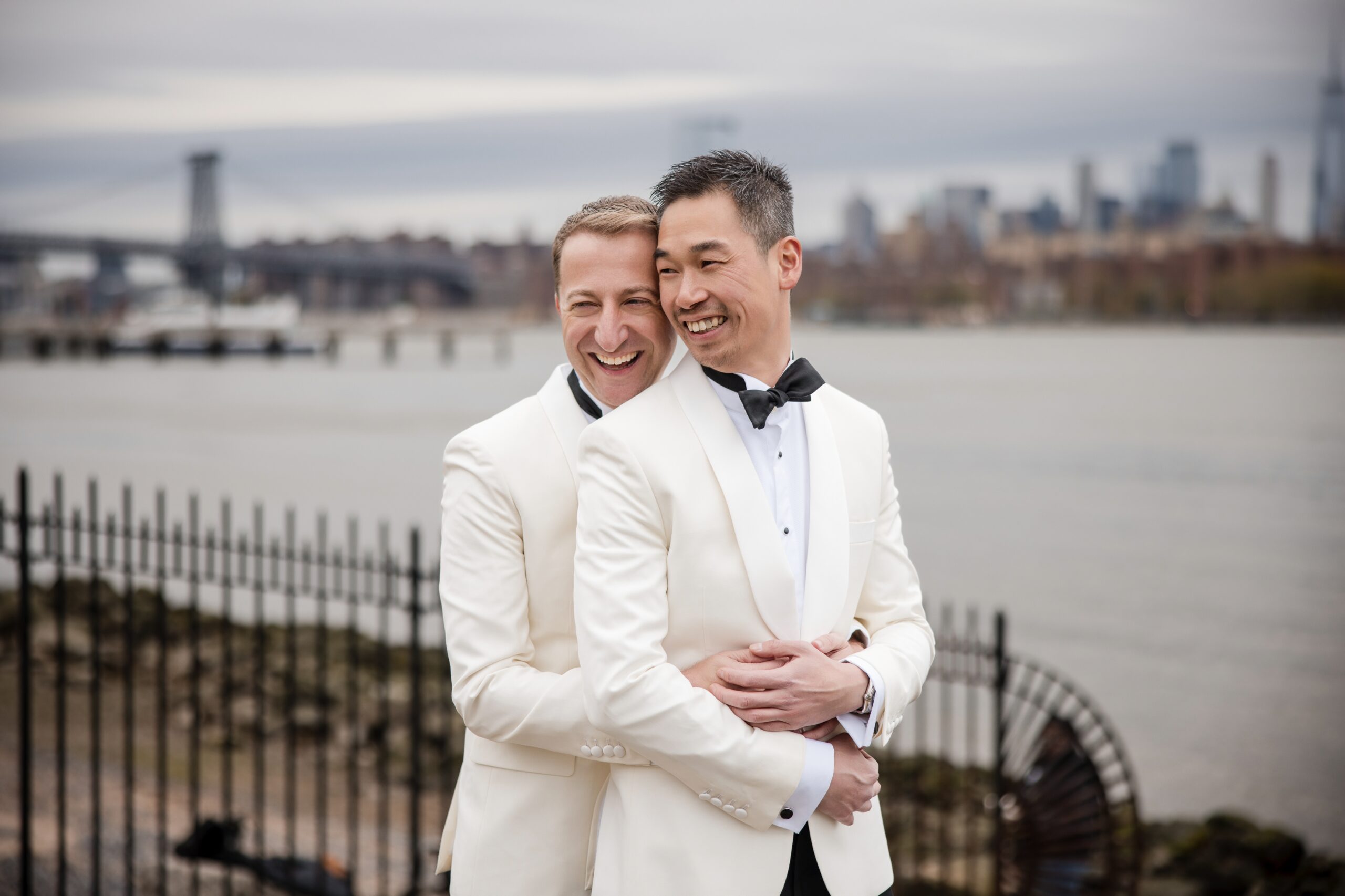 Two grooms in tuxedos embrace, smiling, with a city skyline and river in the background. Two grooms in tuxedos embrace, smiling, with a city skyline and river in the background.