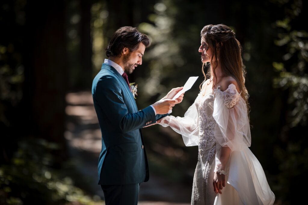 A groom in a blue suit reads from a paper while holding hands with the bride in a white dress at their enchanting Nestldown wedding in a forest setting.