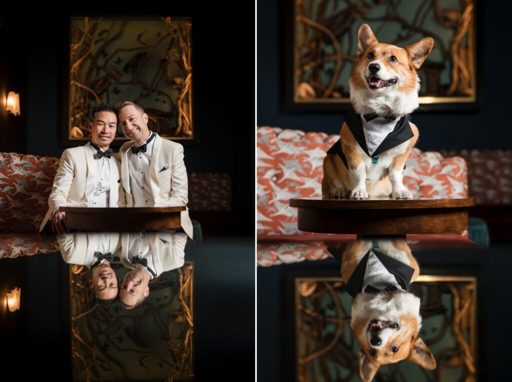 A portrait of a joyful couple in elegant white tuxedos at their 74 Wythe rooftop wedding, seated and reflecting their smiles on a shiny table surface in a warmly lit, artistic room.