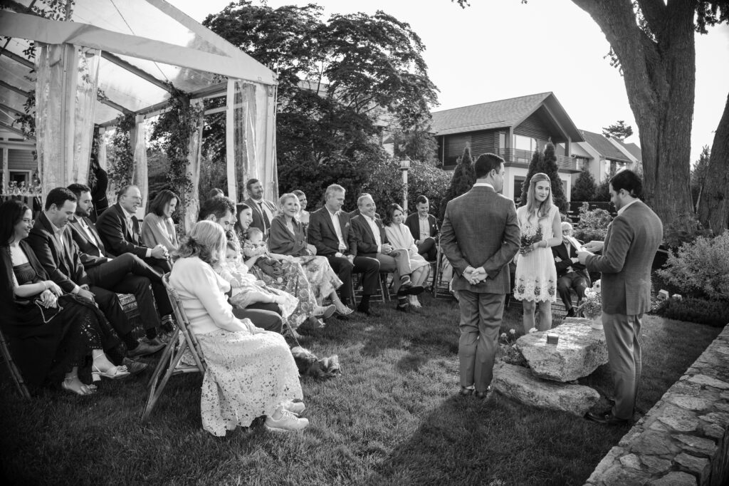 A black and white photo captures an intimate Westport Connecticut wedding. The small outdoor ceremony features a few rows of seated guests, as the couple stands facing the officiant near a stone structure. Trees and houses are visible in the background, adding to the quaint charm of the setting.