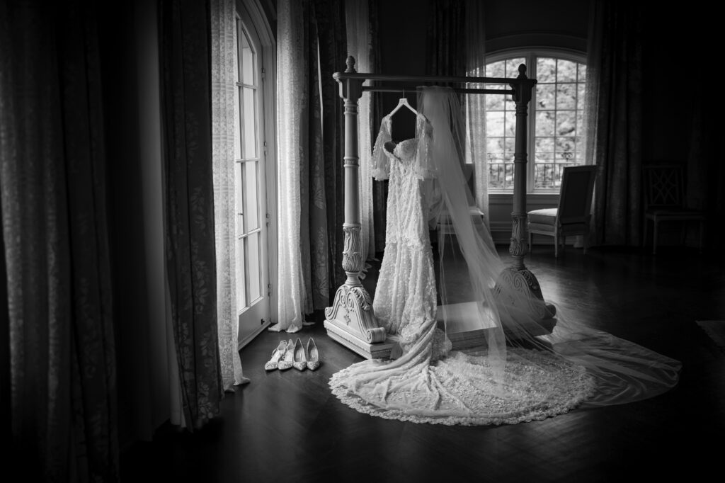 A Park Chateau wedding dress and veil hang on a stand near a window in a dimly lit room. Several pairs of shoes are lined up on the floor next to the stand.