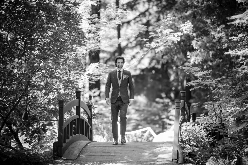 A man in a suit walks across a small wooden bridge in a forested area, perhaps on his way to a Nestldown wedding. The scene is in black and white.
