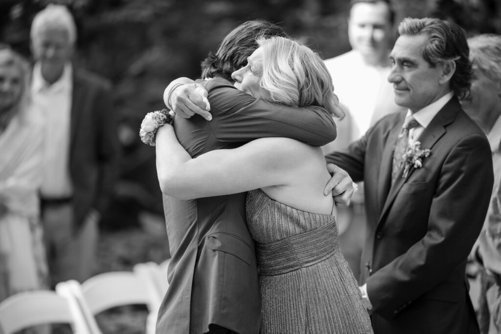 A man and a woman hug at a Nestldown wedding ceremony, with several people standing and observing in the background. The event appears emotional and heartfelt.