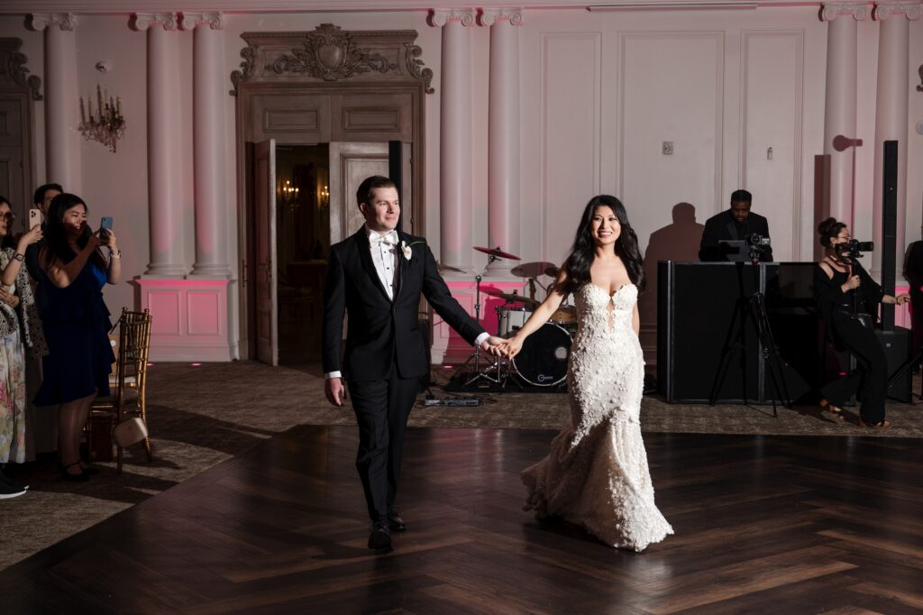 A bride and groom, dressed formally, hold hands and walk together in the beautifully decorated Park Chateau wedding venue while guests watch and take photos.