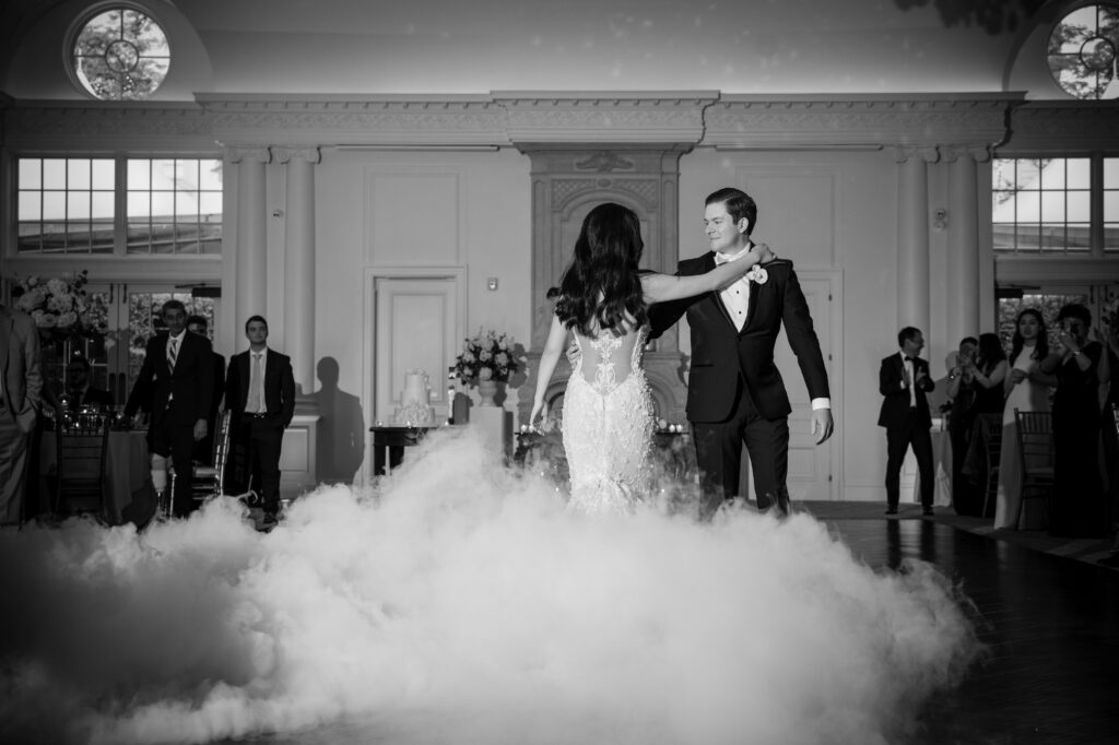A couple dances closely in the center of a room filled with people, with a layer of fog covering the floor, under a bright setting. The scene exudes elegance and formality, capturing the enchanting atmosphere of a Park Chateau wedding.