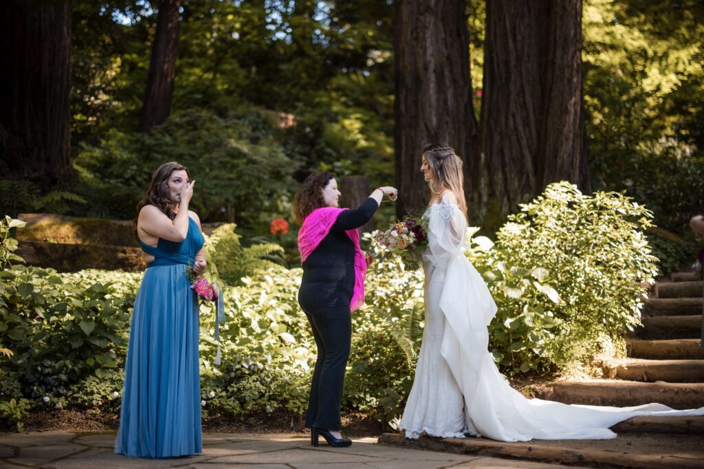 A woman in a wedding dress stands outside at her Nestldown wedding while another woman adjusts her veil and a third woman in a blue dress watches, holding a hand to her face.