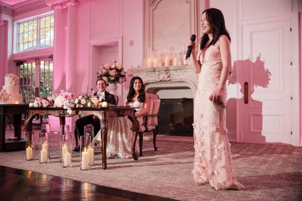 A woman in a floral dress speaks into a microphone at an indoor Park Chateau wedding. Two seated people, a man and a woman, listen attentively. The room is adorned with candles and flowers.