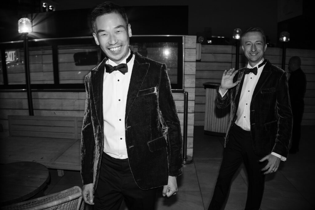 A black and white photograph capturing a joyful moment at a 74 Wythe rooftop wedding, featuring two partners in elegant velvet tuxedos, smiling as they walk through the venue. One partner waves at the camera, adding a lively touch to the scene. 