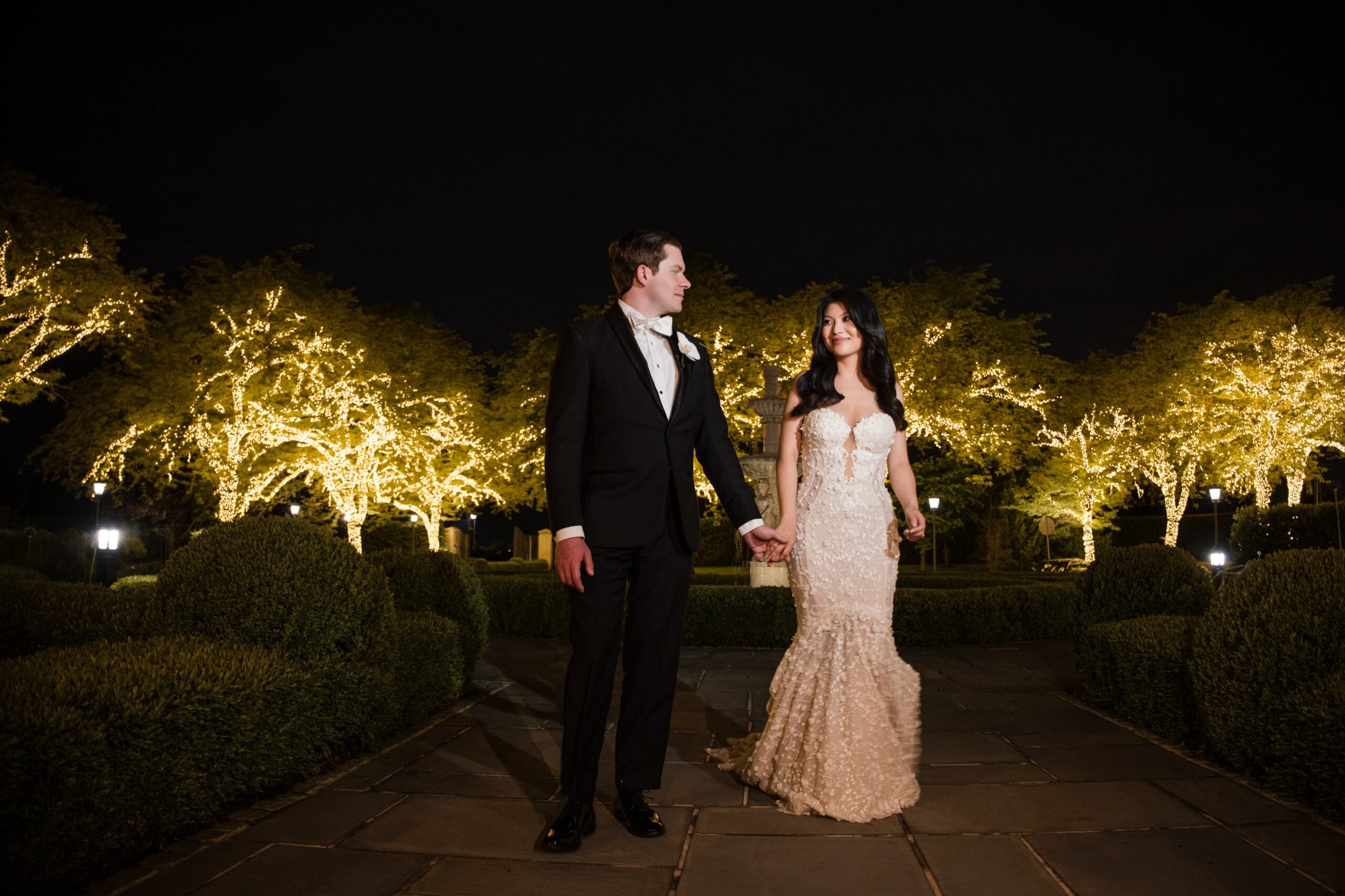 A couple in formal attire walks hand in hand on a stone path at night, with trees decorated with string lights in the background, creating a magical Park Chateau wedding ambiance.