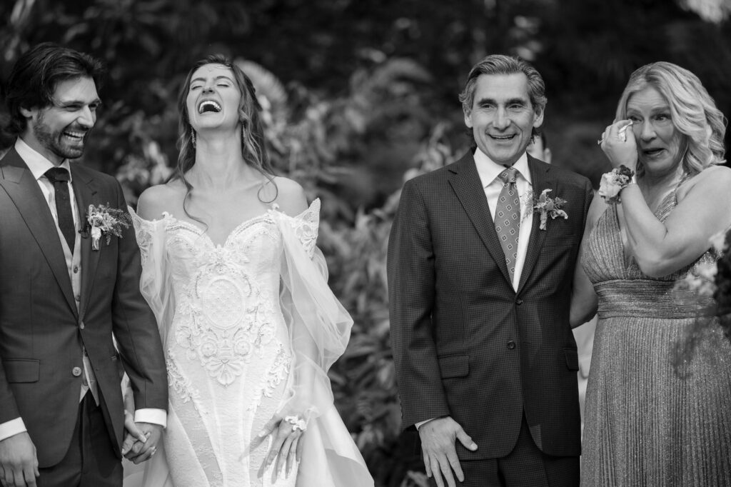 A bride and groom laugh joyously while standing beside two emotionally moved older adults during a Nestldown wedding. The group stands outdoors, surrounded by lush foliage in the background.