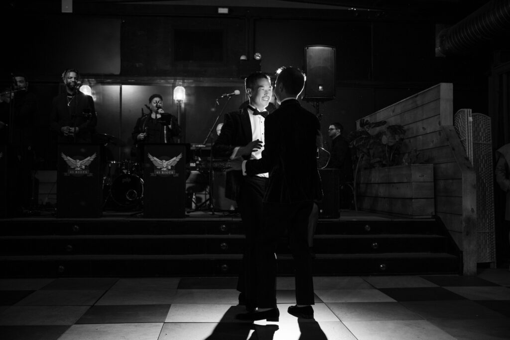 A black and white photograph from a 74 Wythe rooftop wedding, showing a couple sharing their first dance. Lit by a spotlight, they smile at each other, their happiness illuminated against a backdrop of a live band performing in the darkened room. 