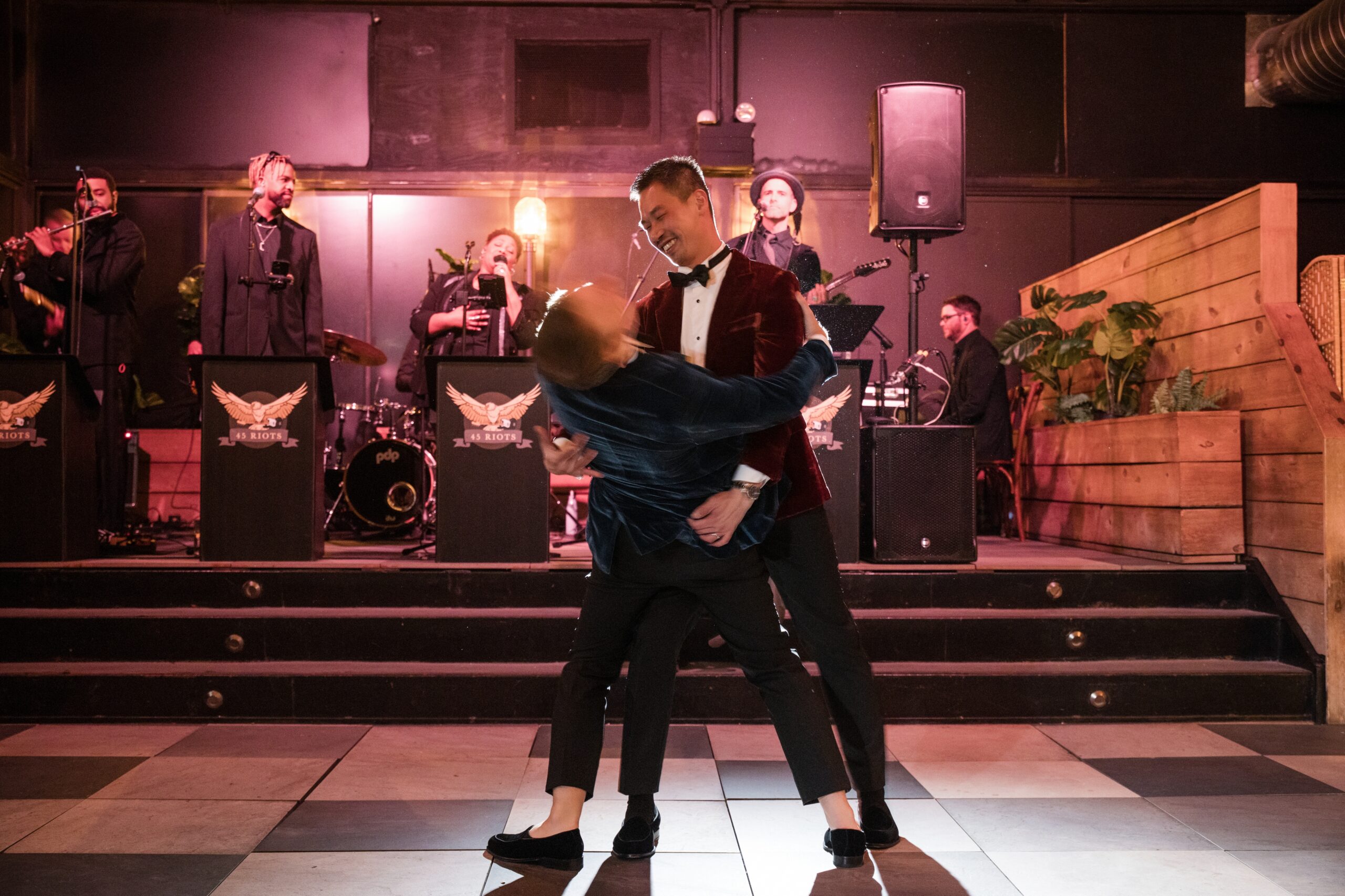 Two grooms dance together on a stage, with one dipping the other dramatically, as a live band performs in the background at their 74 Wythe wedding.