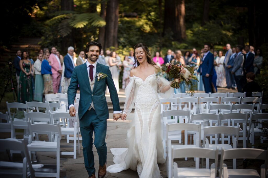 A bride and groom, smiling and holding hands, walk down an aisle outdoors at their Nestldown wedding. Guests in colorful attire stand and watch in the background.