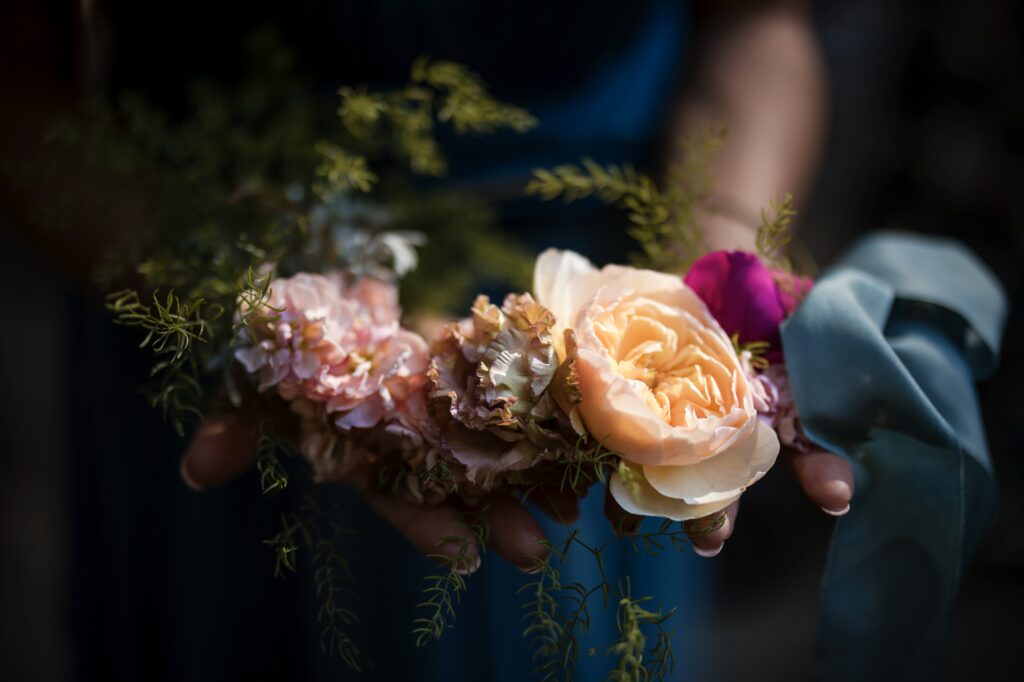 Hands holding a floral arrangement consisting of various flowers, including a cream-colored rose, pink blossoms, and greenery, with a blue ribbon tied around the stems—a perfect bouquet for a Nestldown wedding.