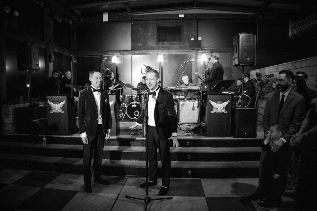 A black and white photograph capturing a heartfelt moment at a 74 Wythe rooftop wedding, where the couple stands at a microphone, addressing their guests. Both dressed in elegant black velvet tuxedos, one partner speaks while the other looks on with a smile, with a live band and attentive guests in the background. 