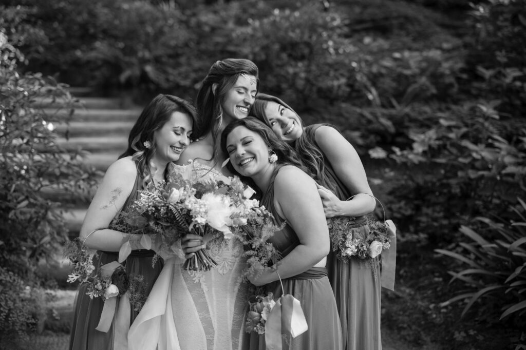 A bride and three bridesmaids, all smiling and laughing, embrace while holding bouquets in the enchanting Nestldown wedding garden.