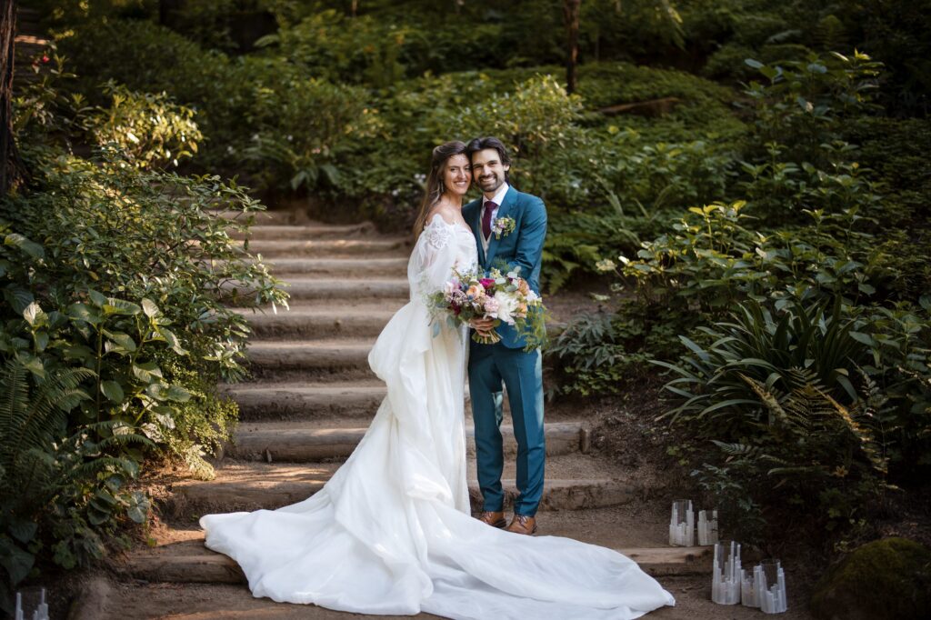 A bride in a white dress and a groom in a blue suit stand together holding a bouquet, smiling on the stone staircase at their Nestldown wedding, surrounded by lush greenery.